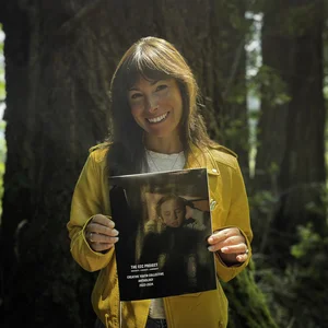 Eine lächelnde Person in gelber Jacke steht in einem Wald und hält ein Buch hoch.