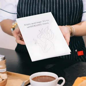 Hands holding book with a white illustrated cover over a coffee cup on a striped placemat in a cafe.