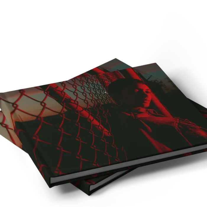 A stack of two landscape, hardcover photo books. The cover shows a dark photo with a red filter, of a person next to a fence