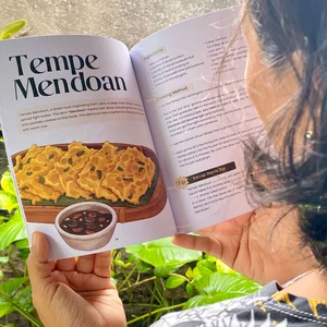 A woman holding the cookbook Tempe and Nasi Campur open on a recipe for Tempe Mendoan with hand drawn illustrations