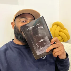 Eine Person mit beigefarbener Mütze hält ein Softcover-Buch mit dem Titel „Sights and Seens“ hoch.