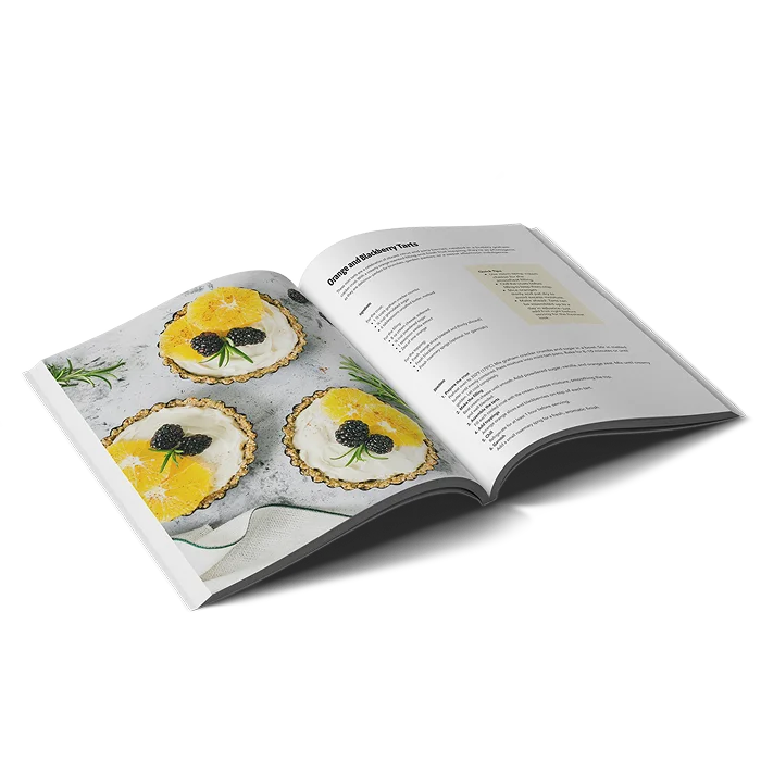 A portrait softcover cookbook open on a recipe for Orange and Blueberry tarts including a photo of three pre-made tarts