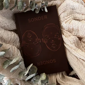 Ein dunkelbraunes Hardcover-Buch mit dem Titel „Sonder“, das auf einer cremefarbenen, gestrickten Oberfläche liegt.